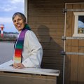 Eine Pastorin steht vor einer kleinen Holzkirche. 
