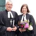 Ein Mann und eine Frau in geistlicher Kleidung (Talar) stehen nebeneinander. Die Frau hält einen Blumenstrauß.